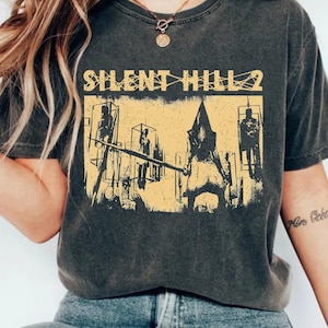 May include: Dark gray t-shirt featuring a distressed yellow graphic from the video game Silent Hill 2. The graphic includes the game's title and a scene with a pyramid-headed figure. The shirt is a vintage style.