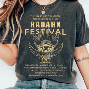 May include: Dark gray t-shirt with gold text and graphic. The text reads "RADAHN FESTIVAL" with a horned creature graphic. Additional text lists champions and locations. The shirt is a vintage style.