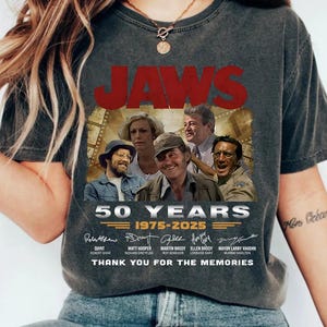 Peut inclure: T-shirt gris foncé avec le titre du film "JAWS" en rouge et une image des acteurs. Le t-shirt commémore les 50 ans du film, avec les années 1975-2025 imprimées en dessous et le texte "THANK YOU FOR THE MEMORIES".