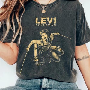 May include: Dark grey t-shirt with a distressed graphic of Levi Ackerman from Attack on Titan. The design includes the name "LEVI ACKERMAN" in a textured font, with Levi in action, holding blades. A casual, vintage-style tee.