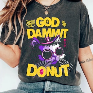 Może przedstawiać: Ciemnoszara koszulka z grafiką przedstawiającą fioletowego kota w okularach i kapeluszu. Tekst na koszulce brzmi "GOD DAMMIT DONUT" w kolorze żółtym, z napisem "DUNGEON CRAWLER CARL" powyżej. Obecna jest również grafika ananasa.