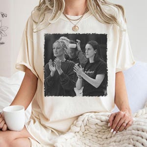 May include: Cream-colored t-shirt featuring a black and white image of two women clapping. One woman wears a black shirt with the word "indiana" on it. The other woman wears a dark sweater. The shirt is worn by a person.