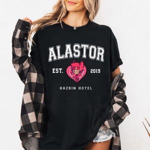 May include: Black t-shirt with white text that reads "ALASTOR EST. 2019 HAZBIN HOTEL". A cartoon character with red horns and a red heart is in the center of the text.