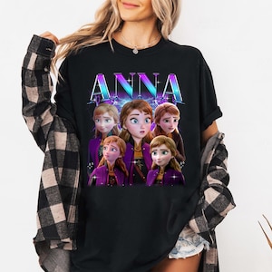 Puede incluir: Camiseta negra con el nombre "ANNA" en una fuente vibrante y degradada. La camiseta muestra un gr&aacute;fico del personaje Anna de la pel&iacute;cula animada, con m&uacute;ltiples im&aacute;genes de ella en varias poses. El dise&ntilde;o tiene un esquema de color p&uacute;rpura y azul.