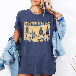 May include: Navy blue t-shirt featuring a distressed yellow graphic from the video game Silent Hill 2. The graphic depicts a scene with figures and the game's title. The shirt is a casual, unisex style.