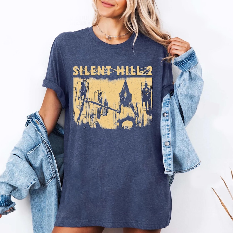 May include: Navy blue t-shirt featuring a distressed yellow graphic from the video game Silent Hill 2. The graphic depicts a scene with figures and the game's title. The shirt is a casual, unisex style.