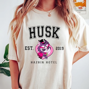 May include: A white t-shirt with a black and pink graphic design. The graphic features a cartoon character with a top hat and the text "Husk Est. 2019 Hazbin Hotel".