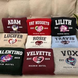 May include: Nine different colored sweatshirts with cartoon character graphics and the text "Hazbin Hotel" on each. The characters include Adam, Fat Nuggets, Lilith, Lucifer, Rosie, Travis, Valentino, Velvette, and Vox.