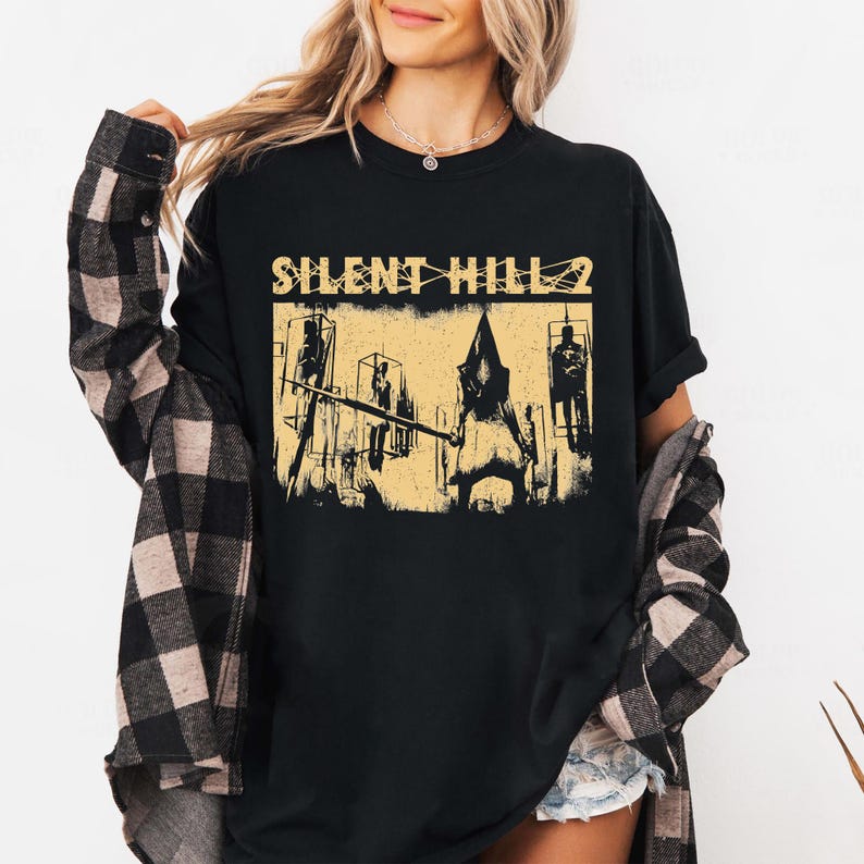May include: Black t-shirt featuring a distressed graphic from Silent Hill 2. The design includes the game's title in a yellow font, along with stylized figures and structures in a similar color. The shirt is a classic crew neck.