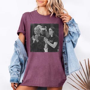 May include: A maroon t-shirt featuring a black and white image of two people clapping. The shirt has short sleeves and a crew neck. The image on the shirt appears to be a photograph. The word "Indiana" is visible on the shirt.