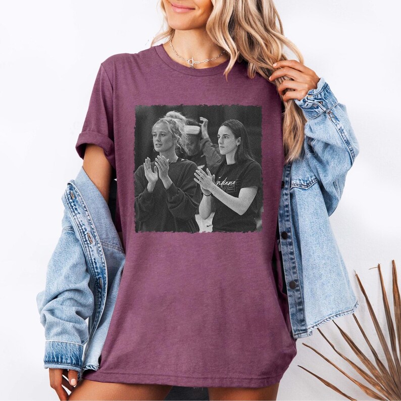 May include: A maroon t-shirt featuring a black and white image of two people clapping. The shirt has short sleeves and a crew neck. The image on the shirt appears to be a photograph. The word "Indiana" is visible on the shirt.