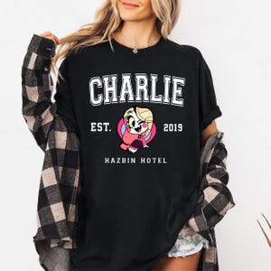 May include: Black t-shirt with white text that reads "CHARLIE EST. 2019 HAZBIN HOTEL". A cartoon character with pink hair and a red dress is in the center of the text.