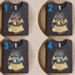 Op de afbeelding: Vier donkergrijze t-shirts met een cartoon drakenontwerp. Elk shirt heeft een draak met een bril en sjaal, zittend op boeken. De sjaals zijn rood, geel, blauw en groen. Er is ook een kleine uil aanwezig.