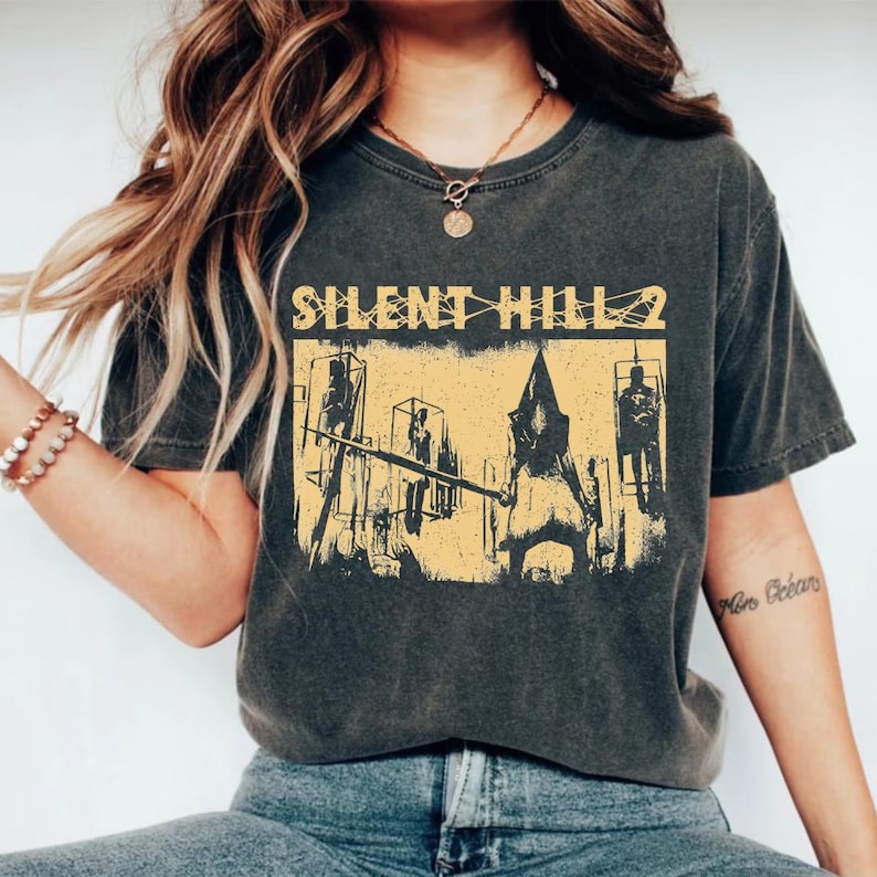 May include: Dark gray t-shirt featuring a distressed yellow graphic from the video game Silent Hill 2. The graphic includes the game's title and a scene with a pyramid-headed figure. The shirt is a vintage style.
