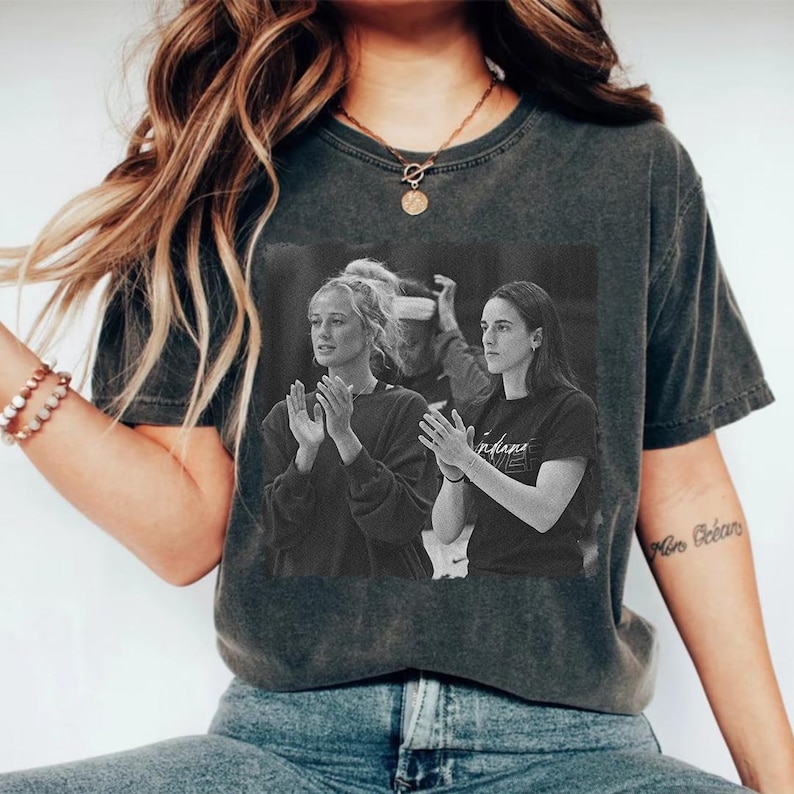 May include: Dark gray t-shirt featuring a black and white image of two women clapping. The shirt has a vintage, worn look. The image on the shirt appears to be a photograph of a sporting event. The shirt is a crew neck.
