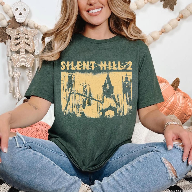 May include: Dark green t-shirt featuring a yellow graphic from Silent Hill 2. The graphic depicts a scene with figures and the text "SILENT HILL 2". The shirt is made of a soft material and is suitable for casual wear.