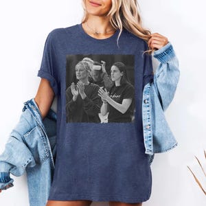 May include: Navy blue t-shirt featuring a black and white image of two women clapping. The image is centered on the shirt. The shirt is worn with a light blue denim jacket.