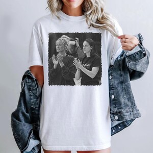 May include: White t-shirt featuring a black and white image of two people clapping. The image is framed with a black border. The person on the right is wearing a black shirt with the word "indiana" printed on it.