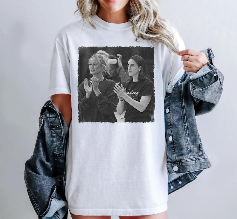 May include: White t-shirt featuring a black and white image of two people clapping. The image is framed with a black border. The person on the right is wearing a black shirt with the word "indiana" printed on it.