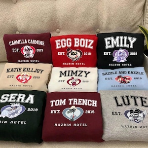 May include: Nine different colored sweatshirts with cartoon character graphics and the text "Hazbin Hotel" printed on each. The characters include Katie Killjoy, Razzle and Dazzle, Mimzy, Tom Trench, Sera, Emily, Egg Boiz, Carmilla Carmine, and Lute.