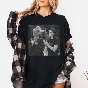 May include: Black t-shirt featuring a grayscale image of two women clapping. The image is framed within a rectangle. The text "Indiana" is visible on the right side of the image. The shirt is worn by a person with long blonde hair.
