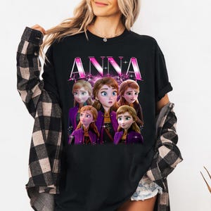 Puede incluir: Camiseta negra con la palabra "ANNA" en rosa, con un gr&aacute;fico de Anna y otros personajes de la pel&iacute;cula Frozen. Los personajes llevan trajes morados y marrones. Cuello redondo.