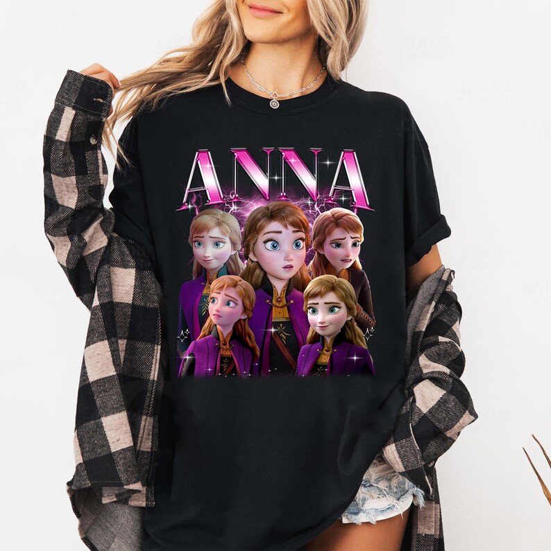Puede incluir: Camiseta negra con la palabra "ANNA" en rosa, con un gr&aacute;fico de Anna y otros personajes de la pel&iacute;cula Frozen. Los personajes llevan trajes morados y marrones. Cuello redondo.