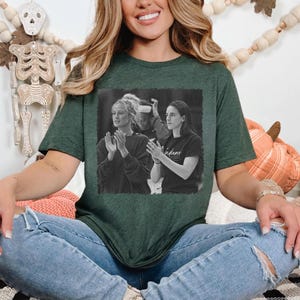 May include: Dark green t-shirt featuring a black and white image of two women clapping. The image is framed in a black rectangle. The t-shirt is worn by a person wearing blue jeans.