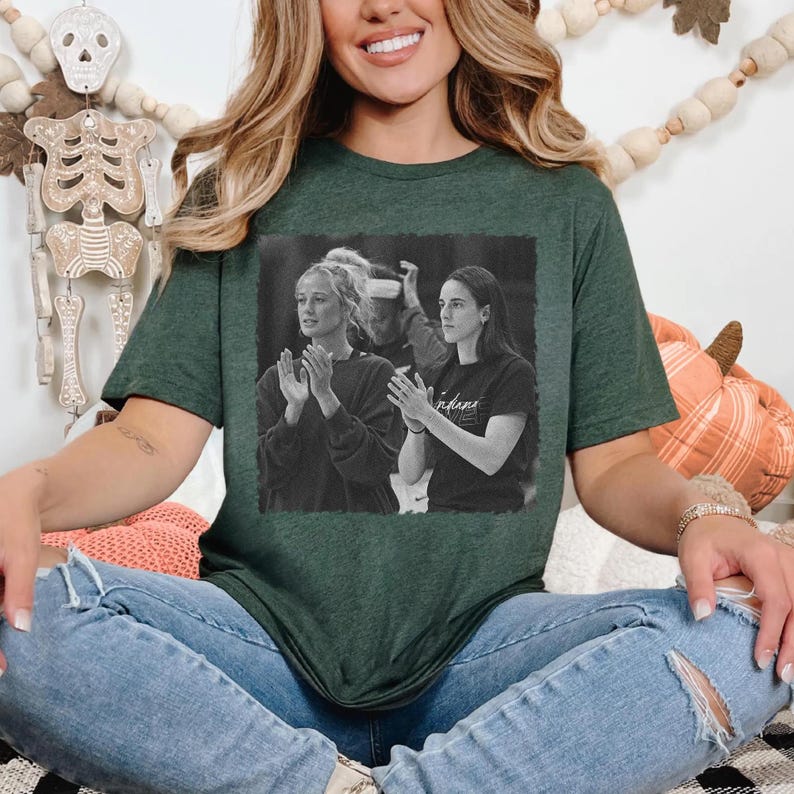 May include: Dark green t-shirt featuring a black and white image of two women clapping. The image is framed in a black rectangle. The t-shirt is worn by a person wearing blue jeans.