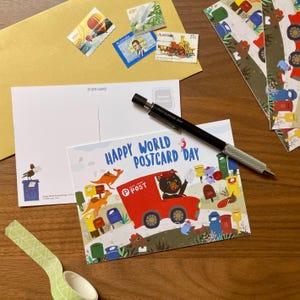 May include: A postcard featuring a red mail truck driven by a bear with the text Happy World Postcard Day, surrounded by kangaroos and mailboxes. A black mechanical pencil rests on top, with a blank postcard, postage stamps, and tape nearby.