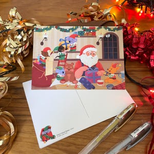 Könnte beinhalten: Eine Weihnachtskarte mit dem Weihnachtsmann und seinen Helfern, darunter Koalas und Bären, die Geschenke in einer Werkstatt verpacken. Die Postkarte ist weiß mit einer farbenfrohen Illustration, zusammen mit goldenen und silbernen Stiften.