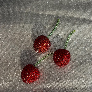 May include: Three red cherry ornaments with green stems, each covered in sparkling red crystals. The cherries are arranged on a shimmering silver surface, creating a festive and eye-catching display.