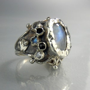 May include: A silver ring with a large, oval-shaped moonstone set in a textured bezel. The ring is adorned with smaller black and white gemstones.