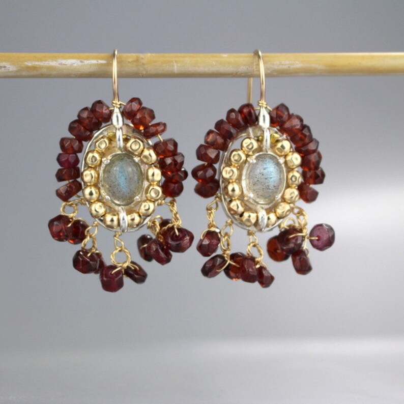 Peut inclure: Une paire de boucles d'oreilles pendantes avec un sertissage argent et or mettant en valeur une pierre de labradorite ronde entour&eacute;e de petites perles dor&eacute;es. Les boucles d'oreilles sont rehauss&eacute;es d'une frange de perles de grenat rouge.
