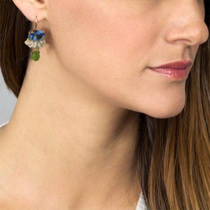 May include: A gold-toned earring with a blue oval gemstone at the top, followed by white and clear stones, and a green teardrop-shaped stone at the bottom. The earring is worn on a person's ear.