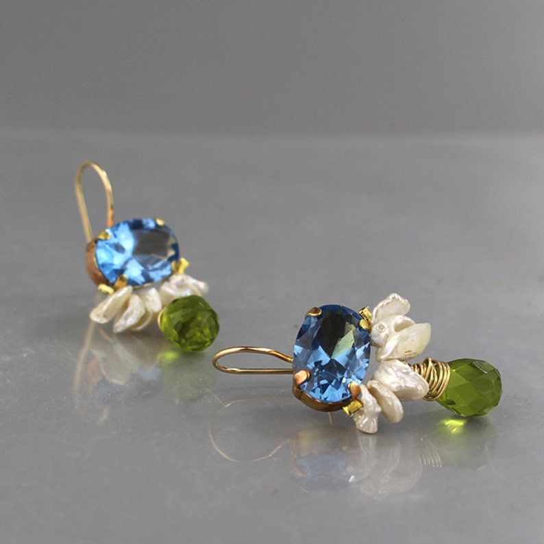 May include: A pair of gold-toned earrings featuring oval, light blue gemstones. Each earring has white floral accents and a green, faceted bead. The earrings are designed with a hook closure.