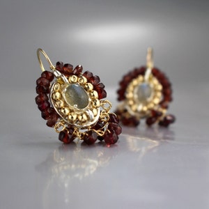 Peut inclure: Une paire de boucles d'oreilles en argent avec un centre dor&eacute; orn&eacute; d'une pierre pr&eacute;cieuse ronde grise entour&eacute;e de petites perles dor&eacute;es. Les boucles d'oreilles sont rehauss&eacute;es de perles de grenat rouge pendantes.