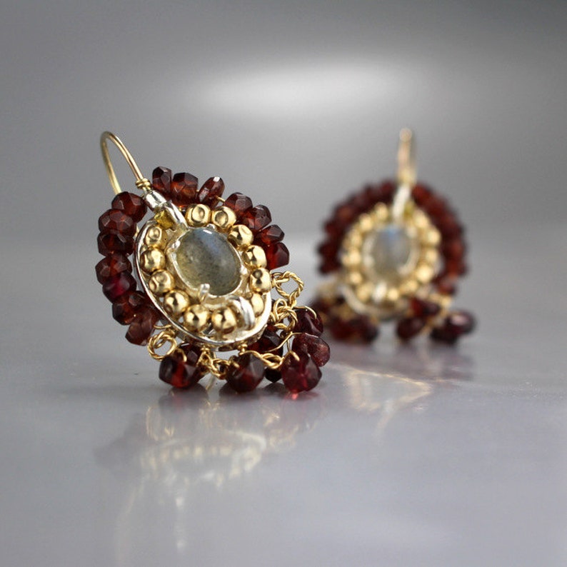 Peut inclure: Une paire de boucles d'oreilles en argent avec un centre dor&eacute; orn&eacute; d'une pierre pr&eacute;cieuse ronde grise entour&eacute;e de petites perles dor&eacute;es. Les boucles d'oreilles sont rehauss&eacute;es de perles de grenat rouge pendantes.