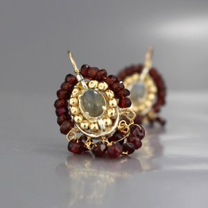 Peut inclure: Boucles d'oreilles en argent et en or avec une pierre pr&eacute;cieuse grise entour&eacute;e de petites perles dor&eacute;es et de perles de grenat rouge.