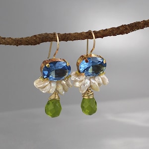 May include: A pair of gold-toned earrings with blue oval-cut gemstones, white pearl clusters, and green teardrop-shaped gems. The earrings are suspended from gold-colored hooks. The jewelry is displayed against a neutral background.