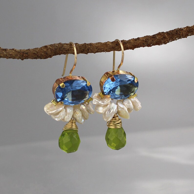 May include: A pair of gold-toned earrings with blue oval-cut gemstones, white pearl clusters, and green teardrop-shaped gems. The earrings are suspended from gold-colored hooks. The jewelry is displayed against a neutral background.