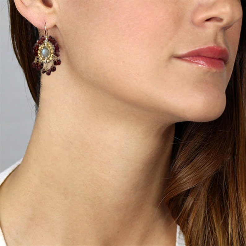 Peut inclure: Une paire de boucles d'oreilles pendantes avec un ton argent&eacute; et dor&eacute;. Les boucles d'oreilles pr&eacute;sentent une pierre pr&eacute;cieuse ronde et iris&eacute;e entour&eacute;e de petites perles dor&eacute;es. La pierre pr&eacute;cieuse est entour&eacute;e d'une frange de petites perles rouges.