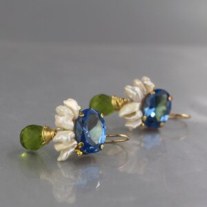 May include: A pair of gold-toned earrings featuring blue oval gemstones, white floral accents, and green teardrop-shaped stones. The earrings are designed with delicate wire wrapping and a hook closure.