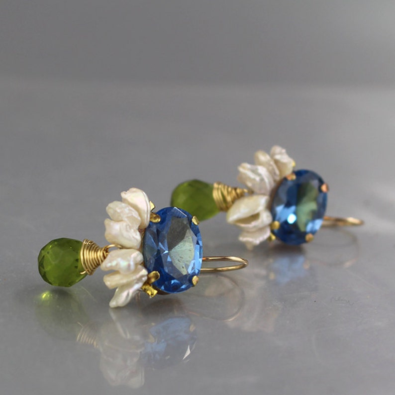 May include: A pair of gold-toned earrings featuring blue oval gemstones, white floral accents, and green teardrop-shaped stones. The earrings are designed with delicate wire wrapping and a hook closure.