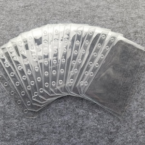 May include: A stack of clear, transparent plastic card sleeves. Each sleeve has a series of small, evenly spaced holes along the edge for binding. The sleeves are rectangular with rounded corners, designed to hold cards.