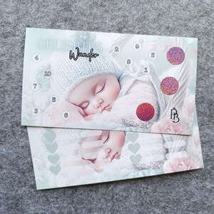 May include: A rectangular card featuring a sleeping baby in a knit hat and sweater. The card displays the text "Wunder" and "GELIEBTES", numbered circles from 1 to 10, and three circular glittery patches in pink and red tones.