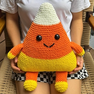 May include: A handmade, crocheted candy corn plush toy. The plush features a white top, orange center, and yellow bottom, with black safety eyes and a smiling face. The toy has arms and legs, and is designed to resemble a candy corn.