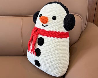Op de afbeelding: Een handgemaakt gehaakt sneeuwman kussen. De sneeuwman heeft een witte body met zwarte knopen, een rode sjaal en zwarte oorbeschermers. Het heeft een wortelneus en zwarte ogen.