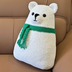 Op de afbeelding: Een handgemaakt wit gehaakt ijsbeerkussen met zwarte ogen en een zwart geborduurde neus. De beer draagt een groene sjaal. Het kussen is ongeveer 38 cm hoog en ligt op een bruin oppervlak.