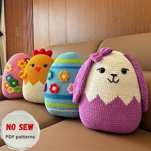 May include: Four colorful, crocheted Easter egg-shaped pillows. The pillows are decorated with flowers, stripes, and animal features. One pillow is shaped like a chick, and another is a bunny. The pillows are on a brown couch.